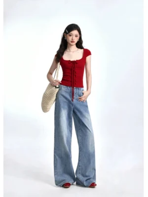 Full body view of a woman wearing a wine red cropped lace-up square neck T-shirt with wide leg blue jeans red flats and woven tote bag
