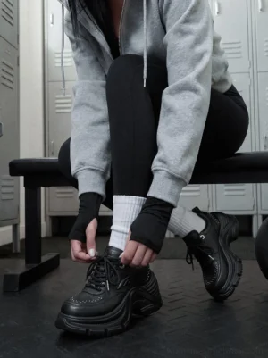 Woman tying black studded chunky platform sneakers in a gym locker room with padded tongue and lace-up streetwear design visible