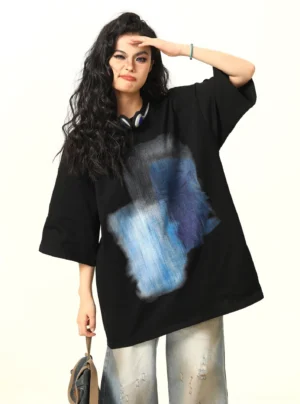 Three-quarter front view of a women's oversized watercolor graphic T-shirt with blue brushstroke print, wide-leg jeans, and shoulder bag