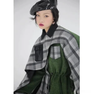 Close-up view of women's asymmetric plaid cape coat showing faux leather pocket trim, black shirt collar, adjustable drawstring waist, and layered green wool-look fabric