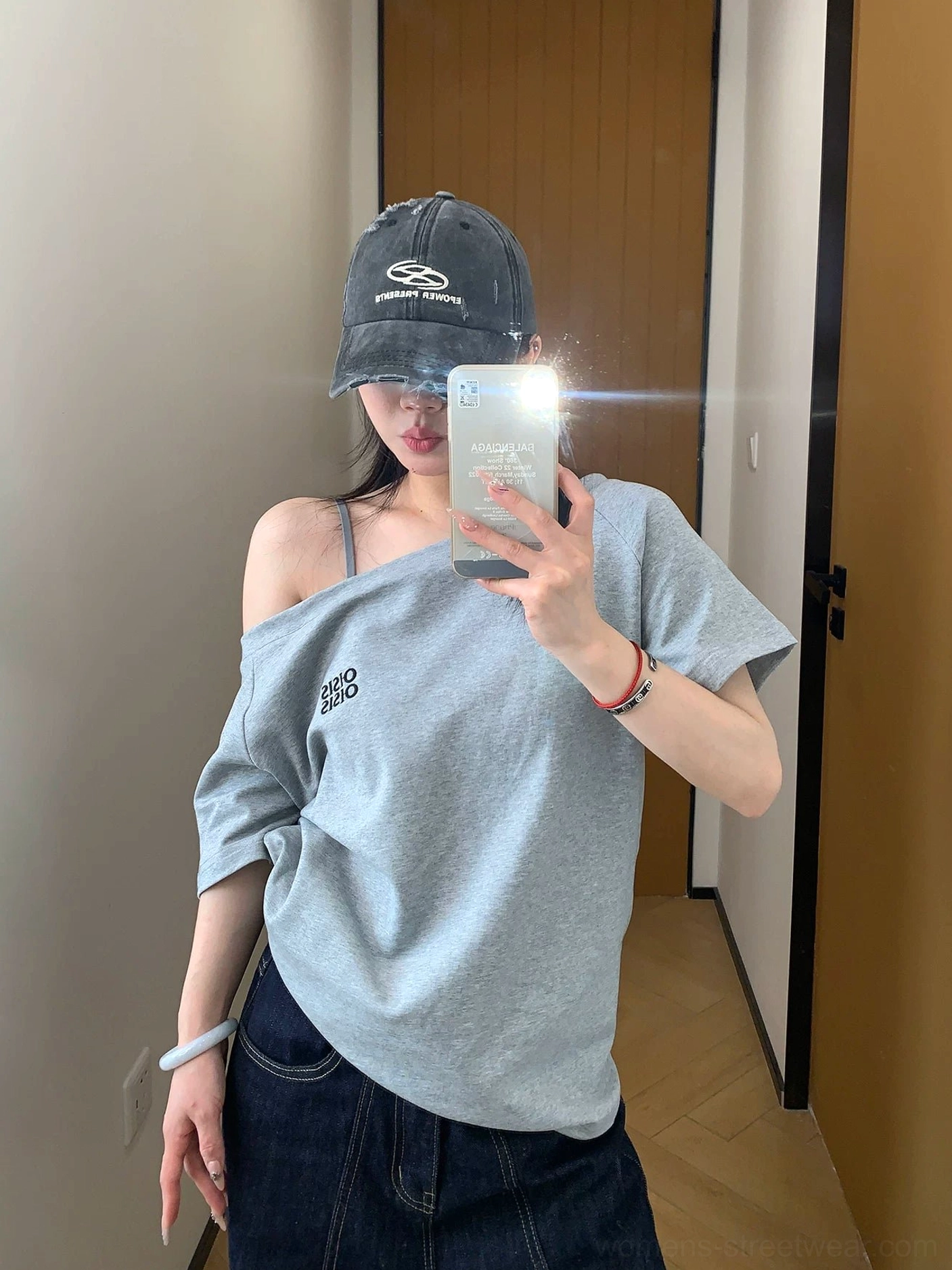 Front mirror view of a heather gray asymmetrical off-shoulder logo tee styled with dark wash jeans and a baseball cap