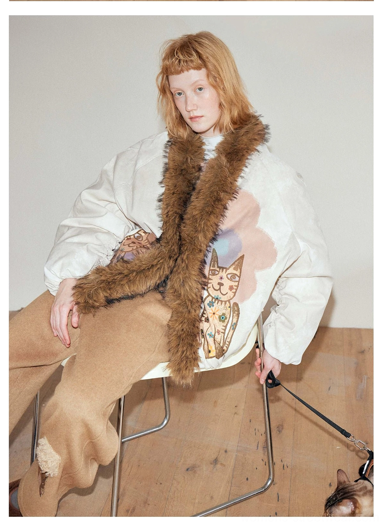 Women's beige faux fur trim bomber jacket with cat and floral artwork shown in a seated streetwear outfit