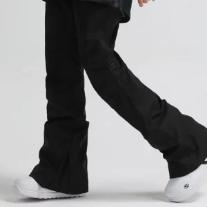 Black women's flared snowboard pants with adjustable waist, side hem zipper, articulated fit, and white ski boots in a low-angle outdoor streetwear outfit
