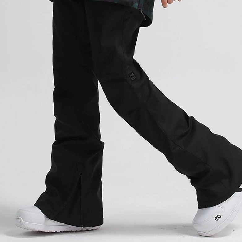 Black women's flared snowboard pants with adjustable waist, side hem zipper, articulated fit, and white ski boots in a low-angle outdoor streetwear outfit