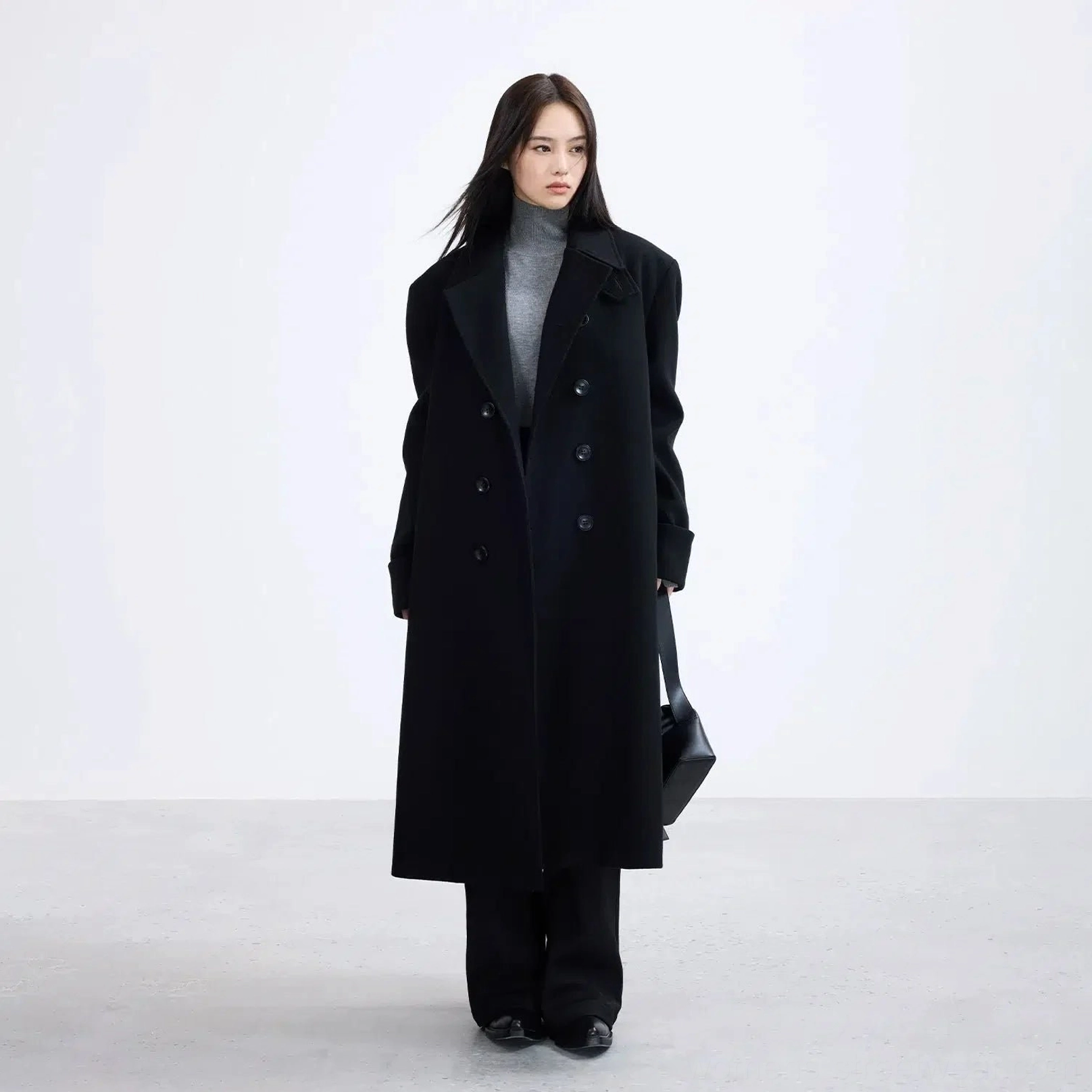 Full-body female model wearing a long black oversized overcoat with turtleneck, suit trousers, and handbag for urban commuting