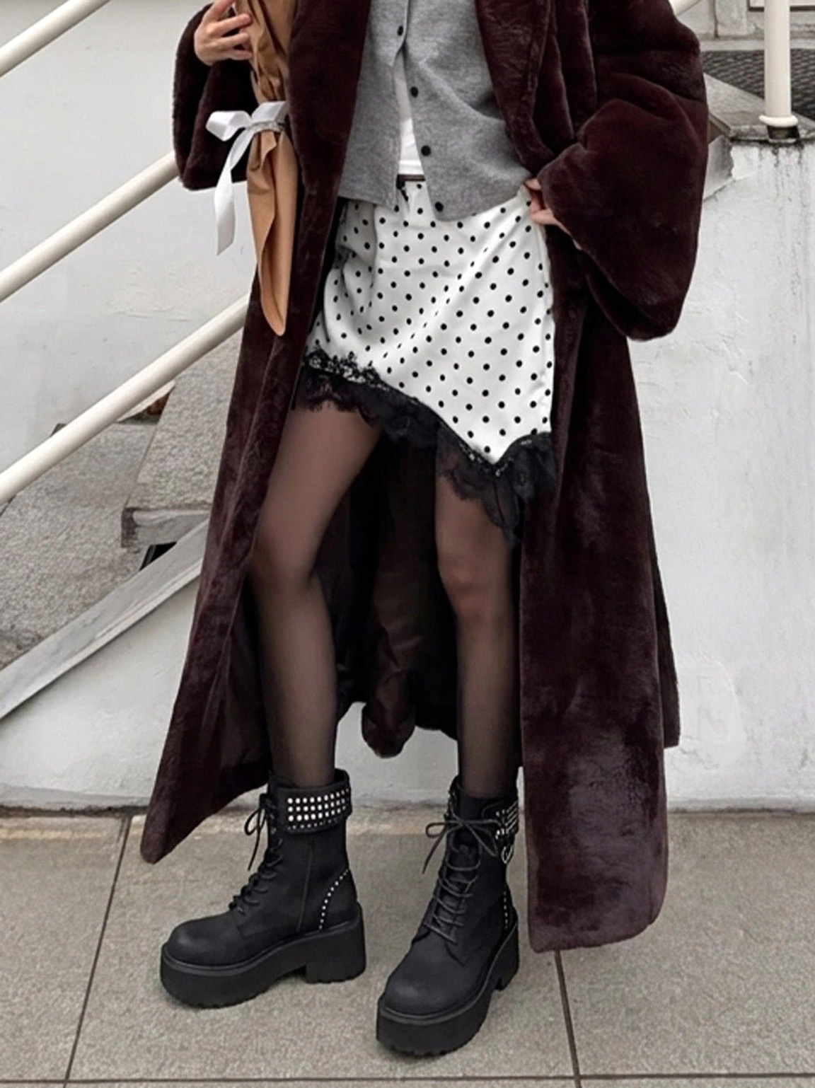 Women's black studded platform combat boots styled with sheer tights, polka dot slip skirt, and long fur coat in a streetwear outfit