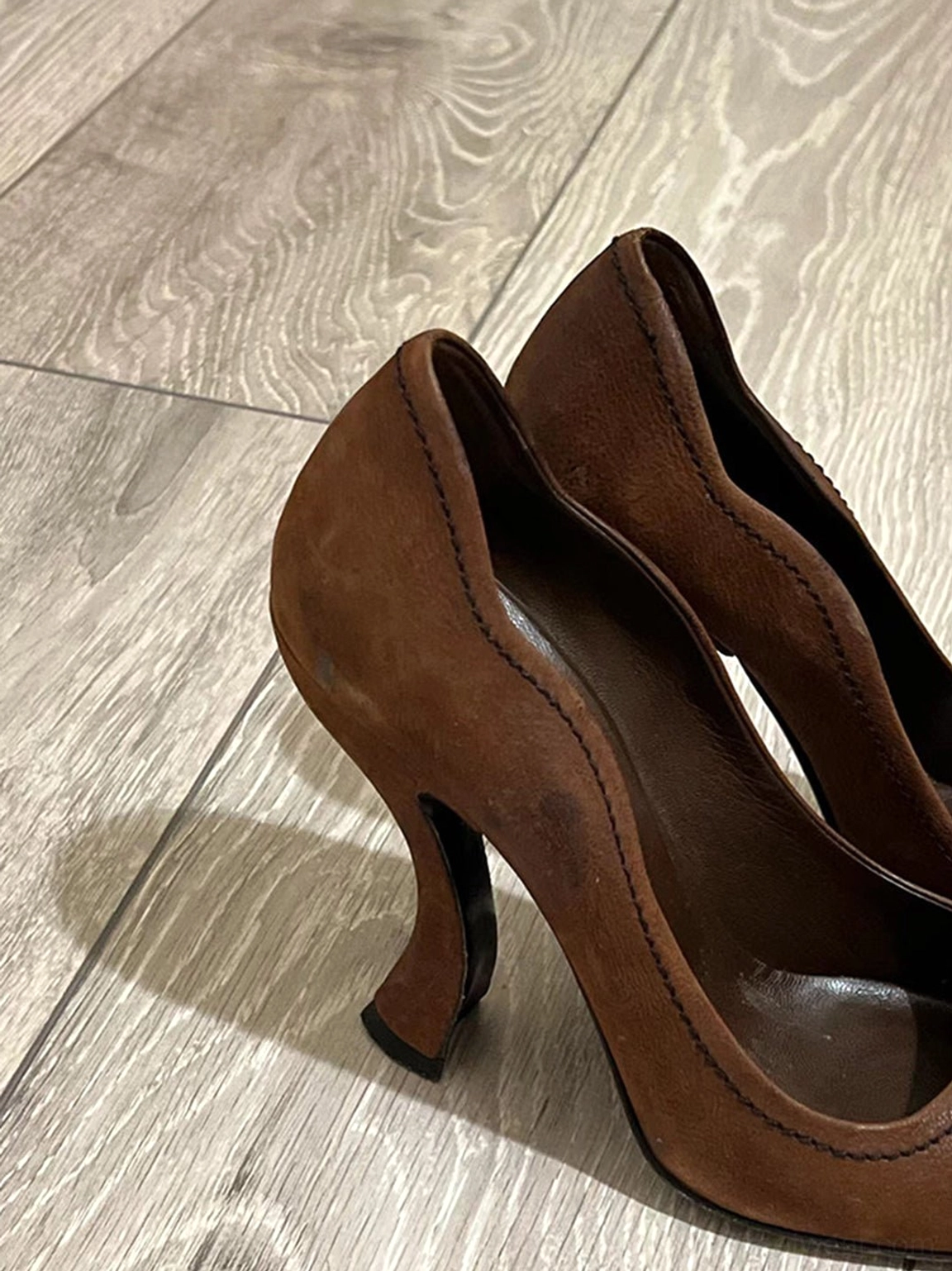 Side three-quarter view of brown round-toe pumps highlighting the curved sculpted high heel and stitched trim