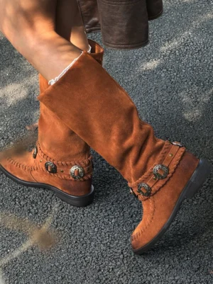 Chestnut brown suede western knee-high boots worn casually on an outdoor road with relaxed slouchy shafts and decorative buckle details
