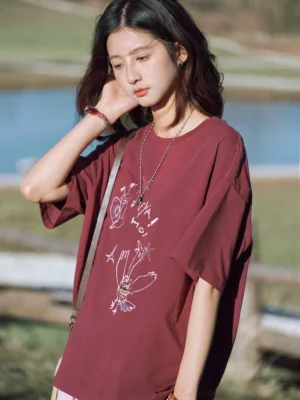 Women's plum color HEYA HO bird sketch oversized graphic T-shirt worn in a lakeside countryside setting