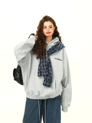 Front three-quarter view of a women’s oversized heather grey plaid accent hoodie with blue plaid hood detail, subtle chest text, wide-leg jeans, and black backpack in a Korean streetwear outfit