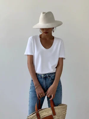 White women's scoop neck cotton T-shirt styled with straight-leg jeans, wide-brimmed hat, and straw tote in a minimalist streetwear outfit