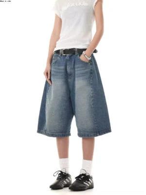 Women's vintage washed denim Bermuda shorts shown from the front with oversized wide legs, low-rise waist, belt styling, white tee, and black retro sneakers