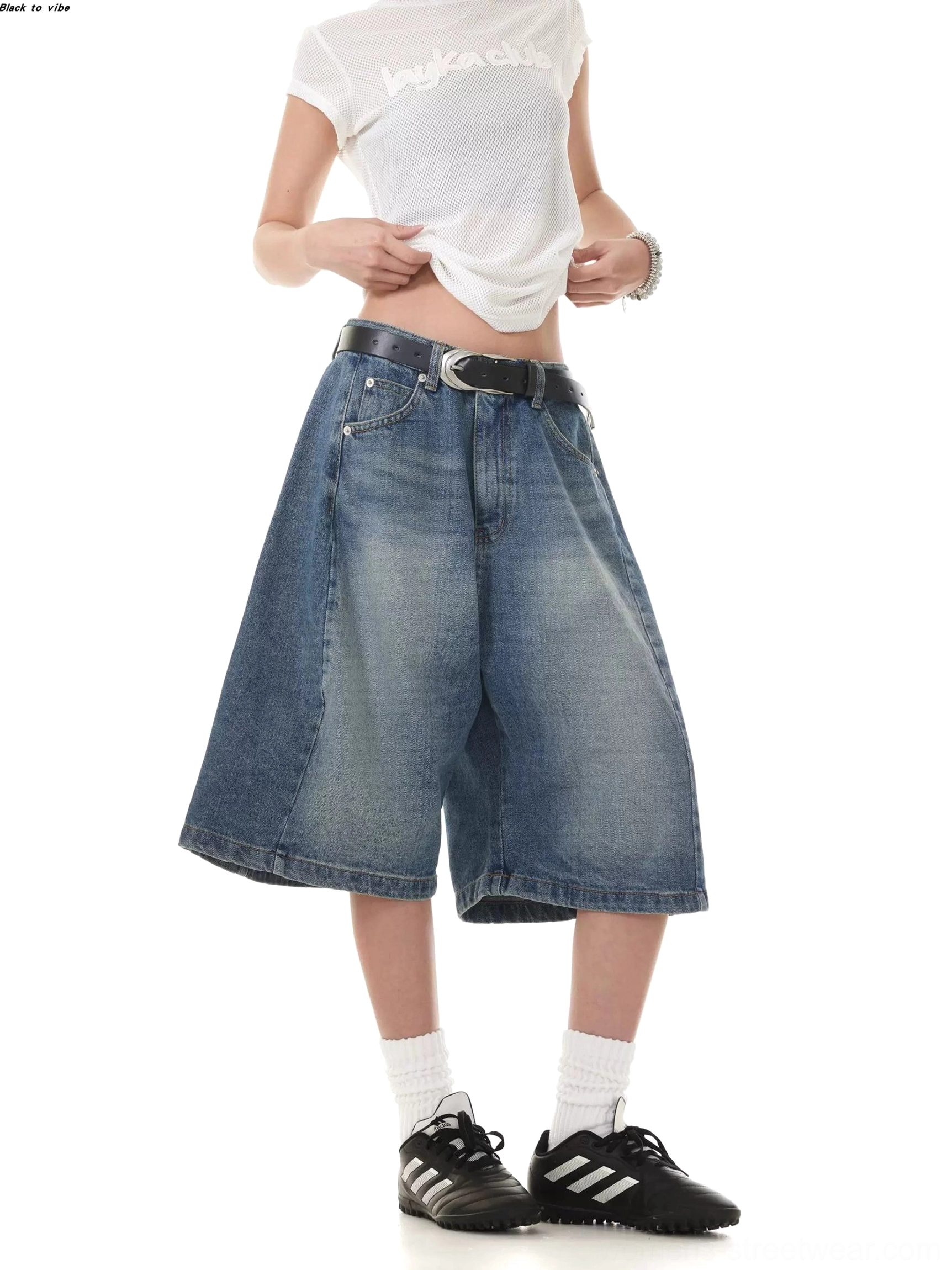 Front view of women's washed blue denim Bermuda shorts with a low-rise waist, relaxed wide-leg fit, vintage fade, cropped white tee, belt, white socks, and black retro sneakers