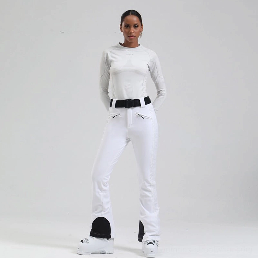 White women's flared ski pants with quilted side panels, black heel guards, zip pockets, and adjustable belt styled with ski boots