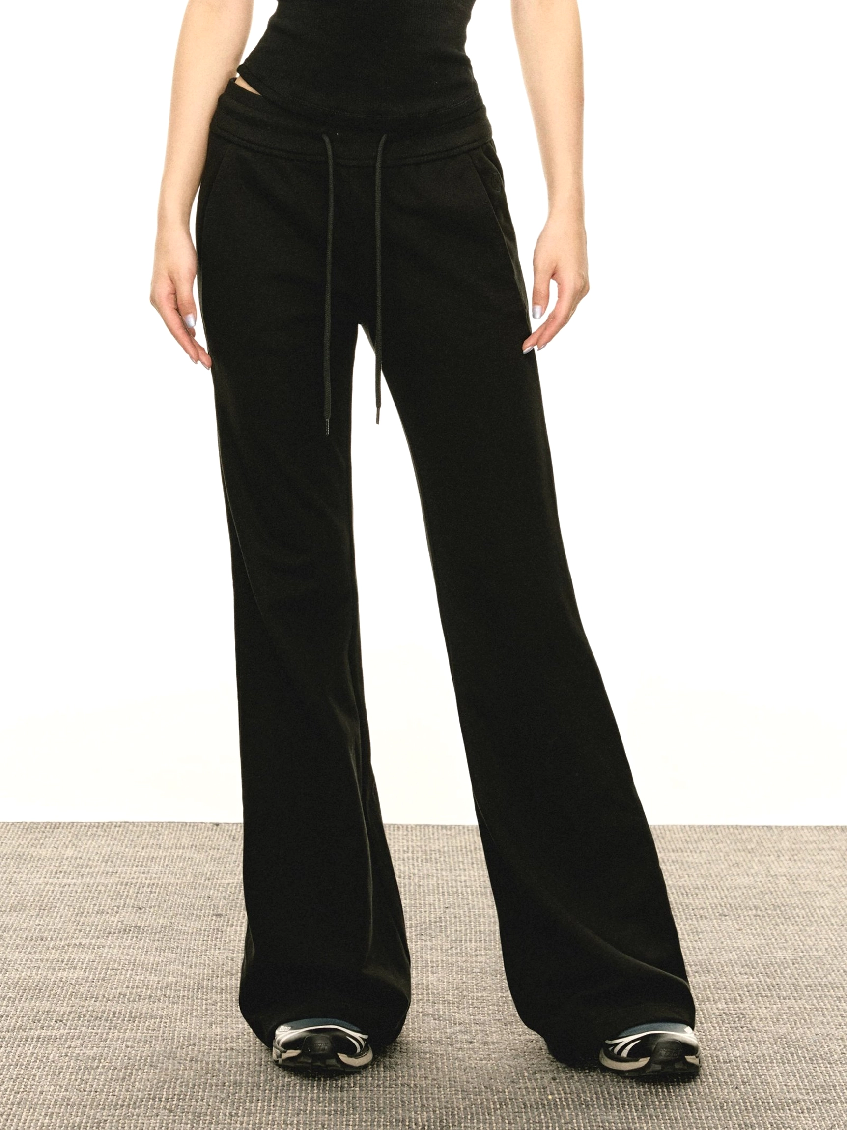 Front full-body view of a model wearing black Y2K flared slim-fit sweatpants with an adjustable drawstring waist, wide-leg silhouette, cropped black top, and sneakers