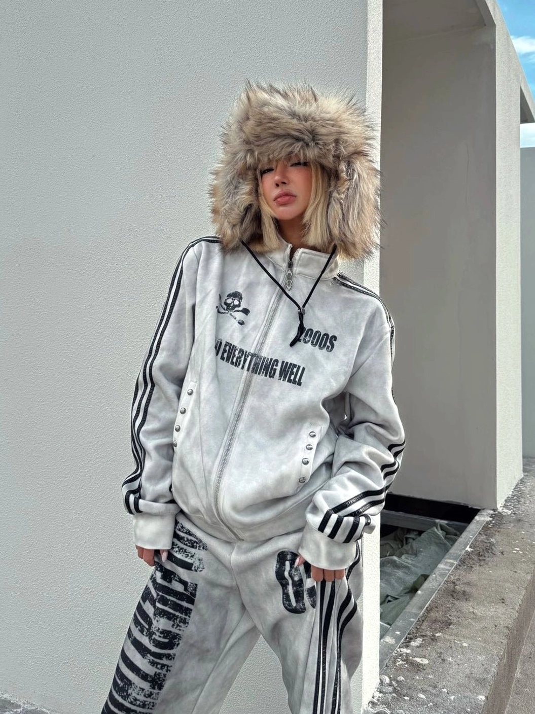 Model wearing a washed white Y2K grunge track jacket with black sleeve stripes, skull graphic, high collar, and matching wide-leg track pants on an industrial rooftop
