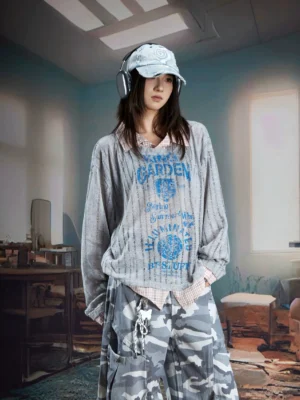Female model wearing a gray sheer layered vintage print shirt sweater with blue graphic artwork, ribbed texture, and built-in plaid shirt details in a retro industrial setting