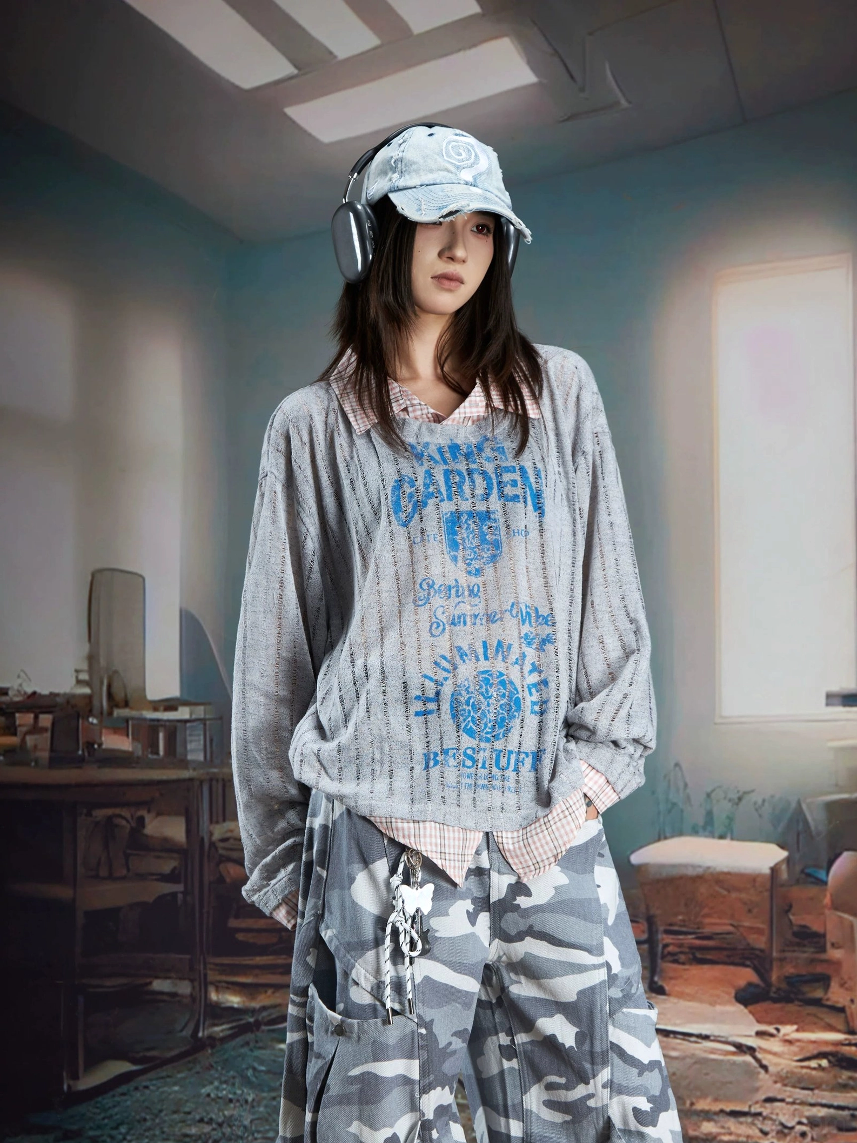 Female model wearing a gray sheer layered vintage print shirt sweater with blue graphic artwork, ribbed texture, and built-in plaid shirt details in a retro industrial setting
