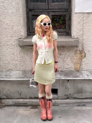 Female model wearing distressed washed pink PU tall boots with tie-dye crop top and beige mini skirt, front full-body outdoor shot against aged concrete building