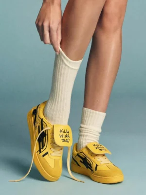 Yellow graffiti retro trainer sneakers worn with white crew socks, showing the low-top silhouette and detachable graphic tongue detail