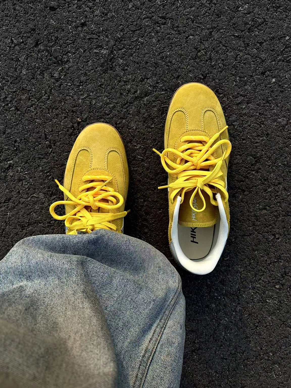 First-person view of yellow lace-up suede trainers worn with blue denim jeans on an urban asphalt street