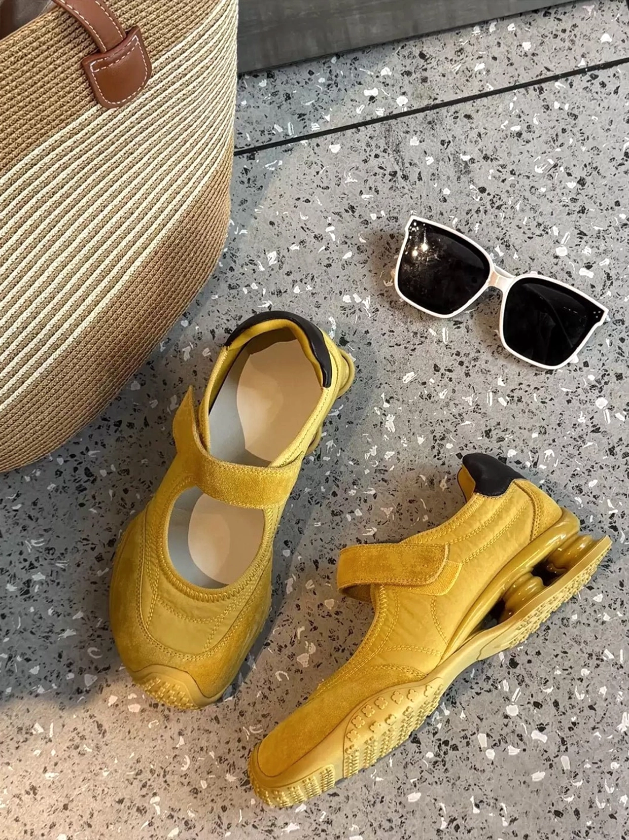 Yellow Mary Jane air cushion ballet trainers flat lay with woven beige tote bag and black square sunglasses on a terrazzo surface