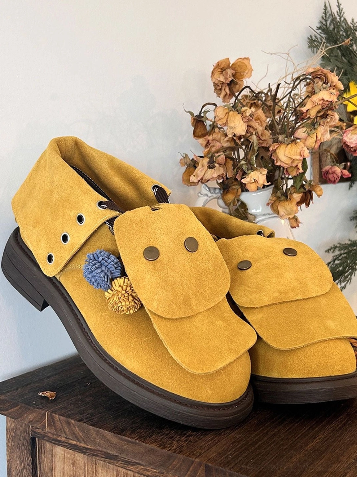 Pair of yellow tassel-flap suede-look ankle boots with blue and yellow braided pom-pom laces displayed on a wooden table