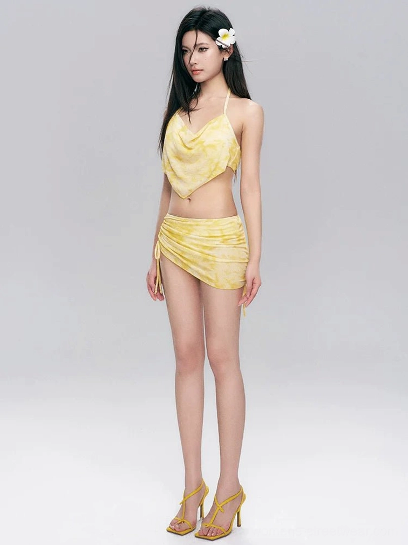 Side-front view of a yellow tie-dye halter bikini and ruched skirt swim set with adjustable side detailing and coordinated yellow high heels